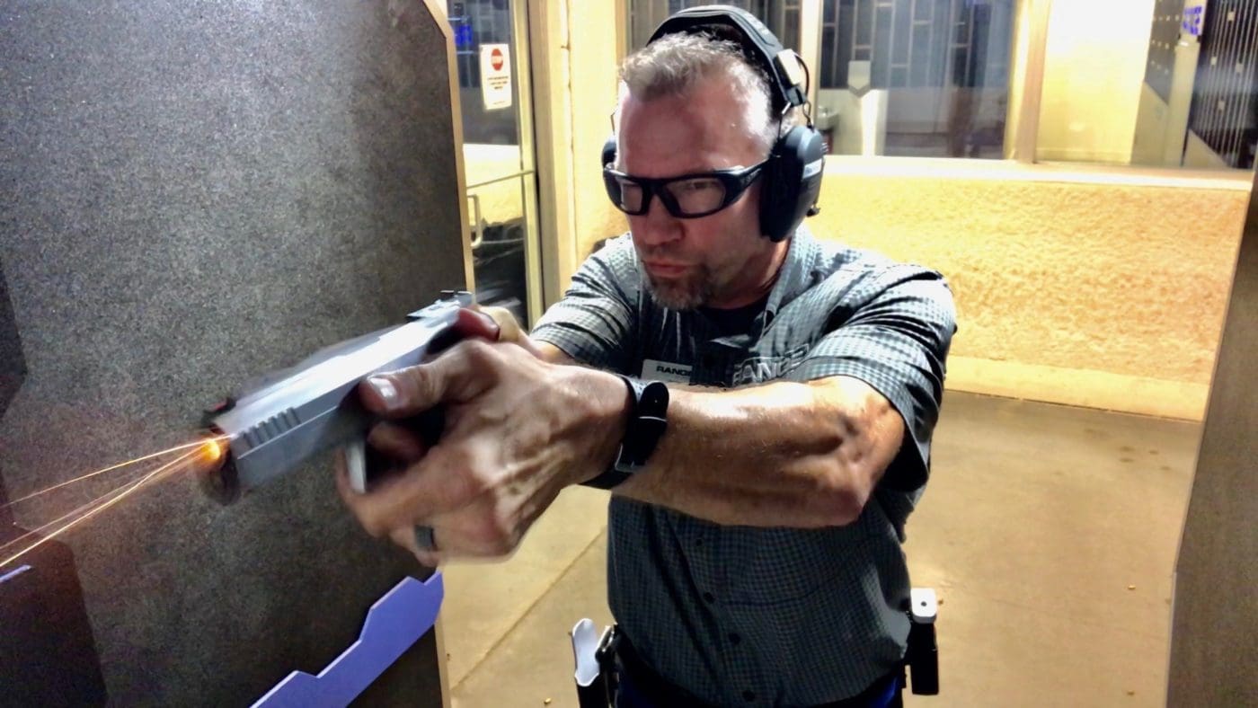 Man wearing ear protection and safety glasses firing a semi-automatic pistol at an indoor shooting range during a live-fire drill.