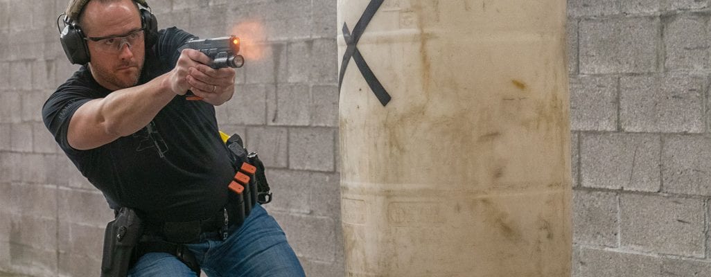 Man in ear and eye protection firing a pistol with a weapon light around a cylindrical barrier at an indoor shooting range, muzzle flash visible.