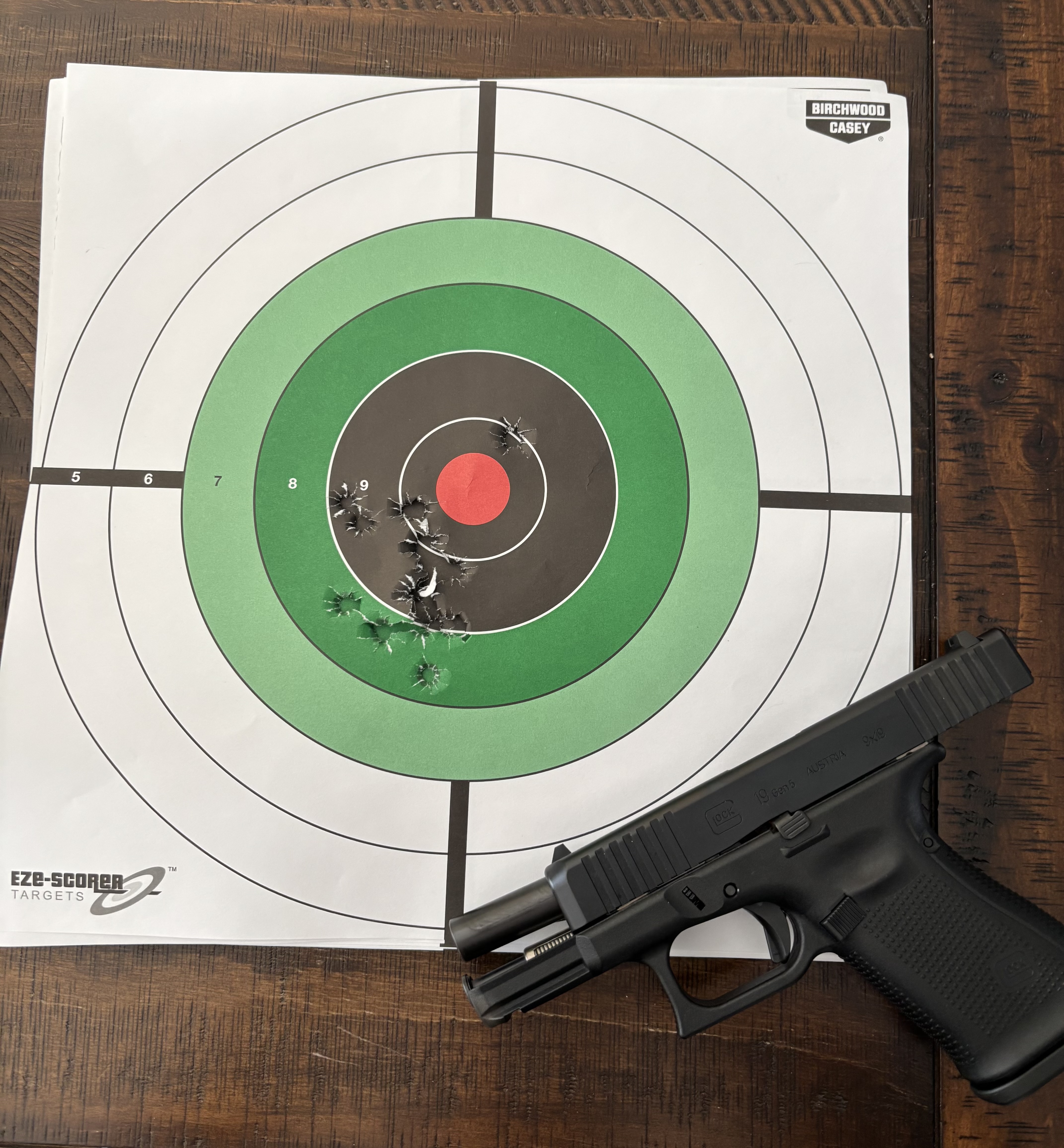 A compact Glock pistol resting beside a paper bullseye target with multiple bullet holes clustered near the center rings on a wooden surface.
