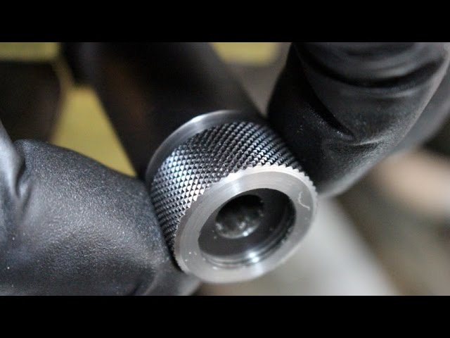 Close-up of a threaded rifle muzzle with a knurled stainless thread protector, held by a gloved hand.