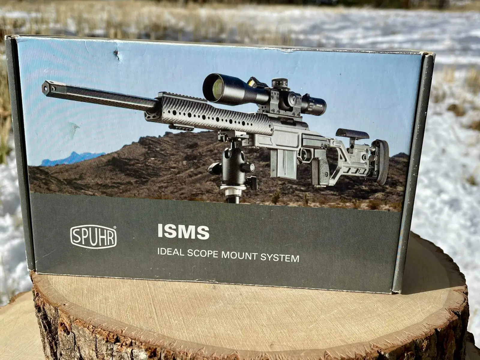 Spuhr ISMS Ideal Scope Mount System retail box resting on a wood stump in a snowy outdoor setting, with product artwork showing a precision bolt-action rifle mounted on a tripod.