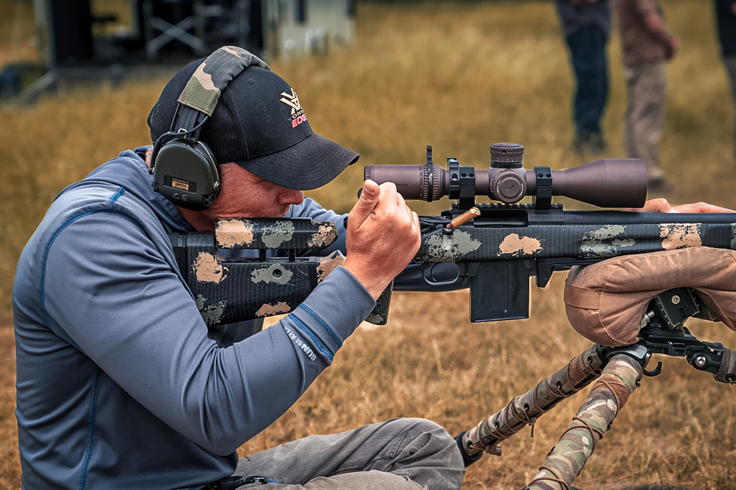 Man in blue pullover shooting a camouflage-patterned carbon fiber chassis bolt-action rifle mounted on a bipod, viewed through a large magnification scope, at an outdoor range.