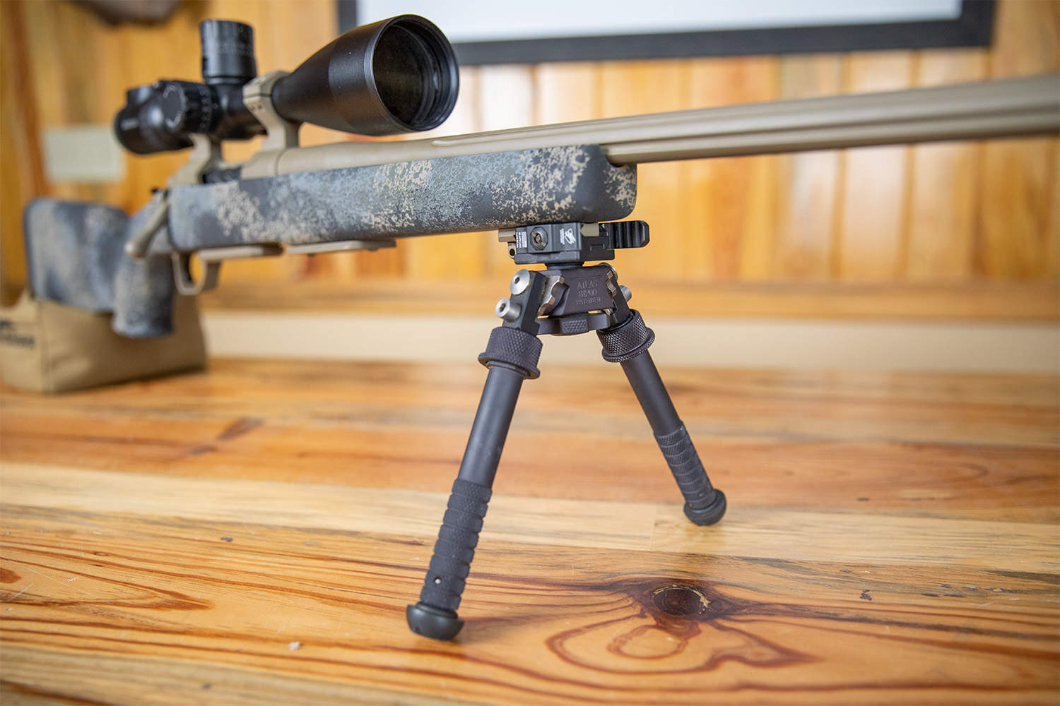 Close-up of an Atlas BT46 bipod attached to a bolt-action precision rifle with a speckled gray stock and large objective scope, resting on a wooden surface.