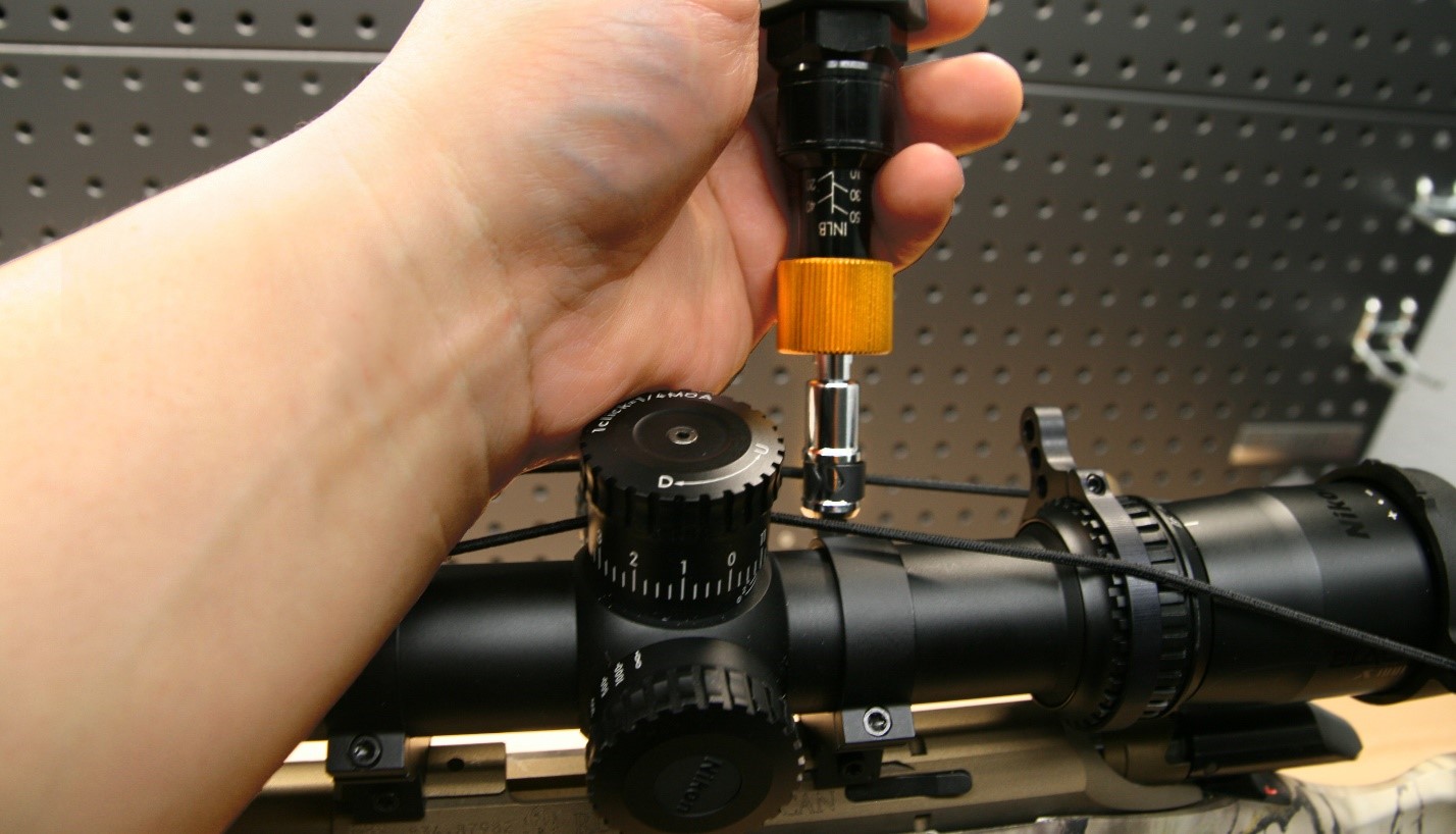 Hand using an inch-pound torque wrench to tighten a scope ring screw on a rifle-mounted riflescope with visible elevation turret markings.
