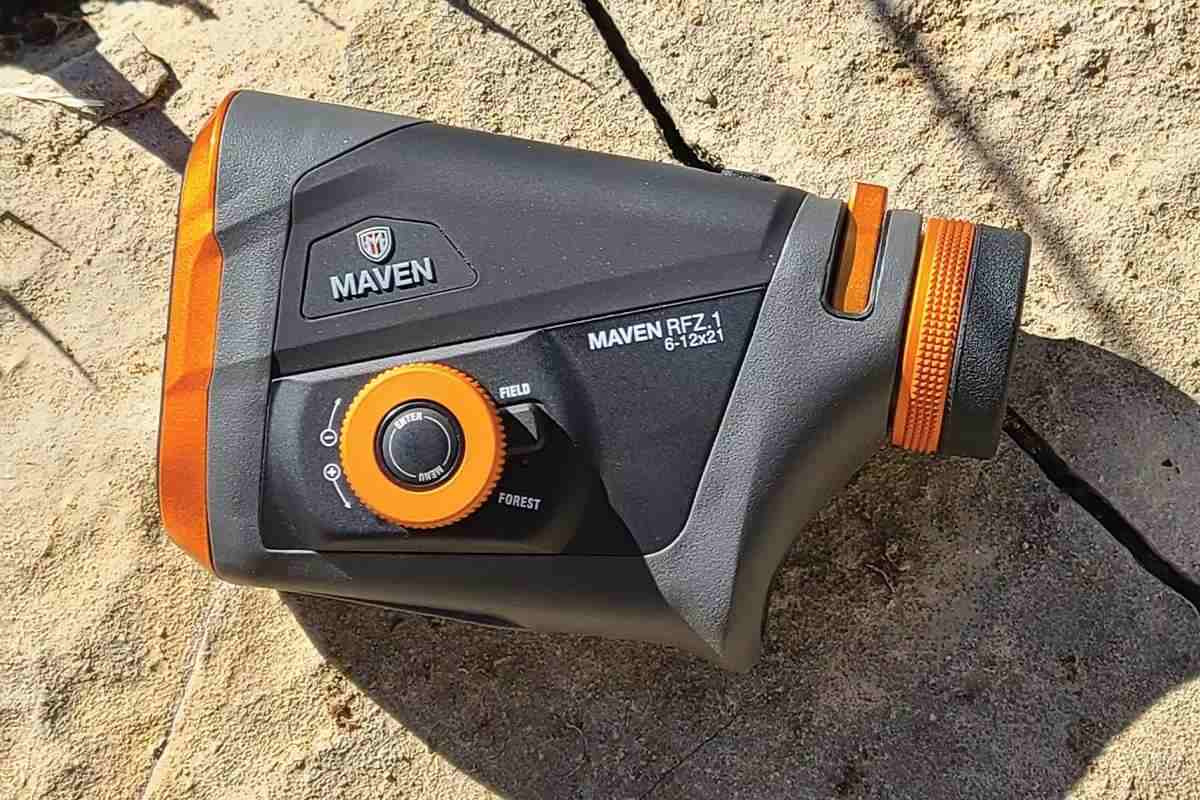 Maven RFZ.1 6-12x21 laser rangefinder in black and orange, showing control dial with Field and Forest mode selector, resting on rocky ground.