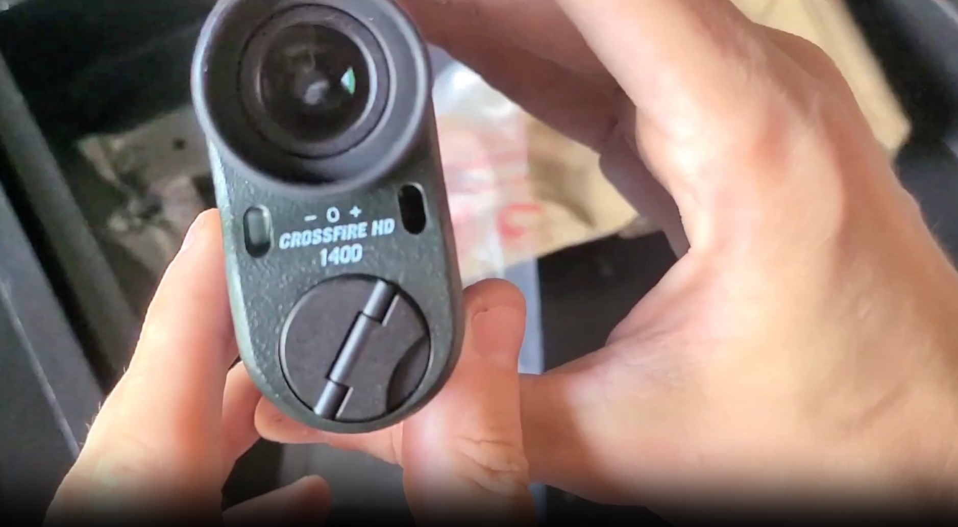 Hands holding a Vortex Crossfire HD 1400 laser rangefinder, showing the objective lens, scan button, and battery compartment cover.