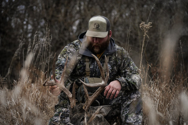 Bearded hunter in camouflage kneeling in tall grass holding the antlers of a harvested whitetail buck.