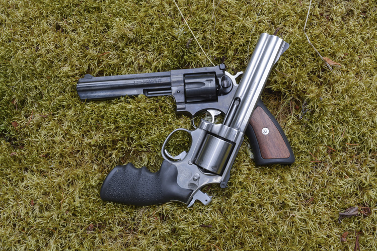 Blued revolver and stainless Ruger GP100 with wood grips crossed on a mossy surface