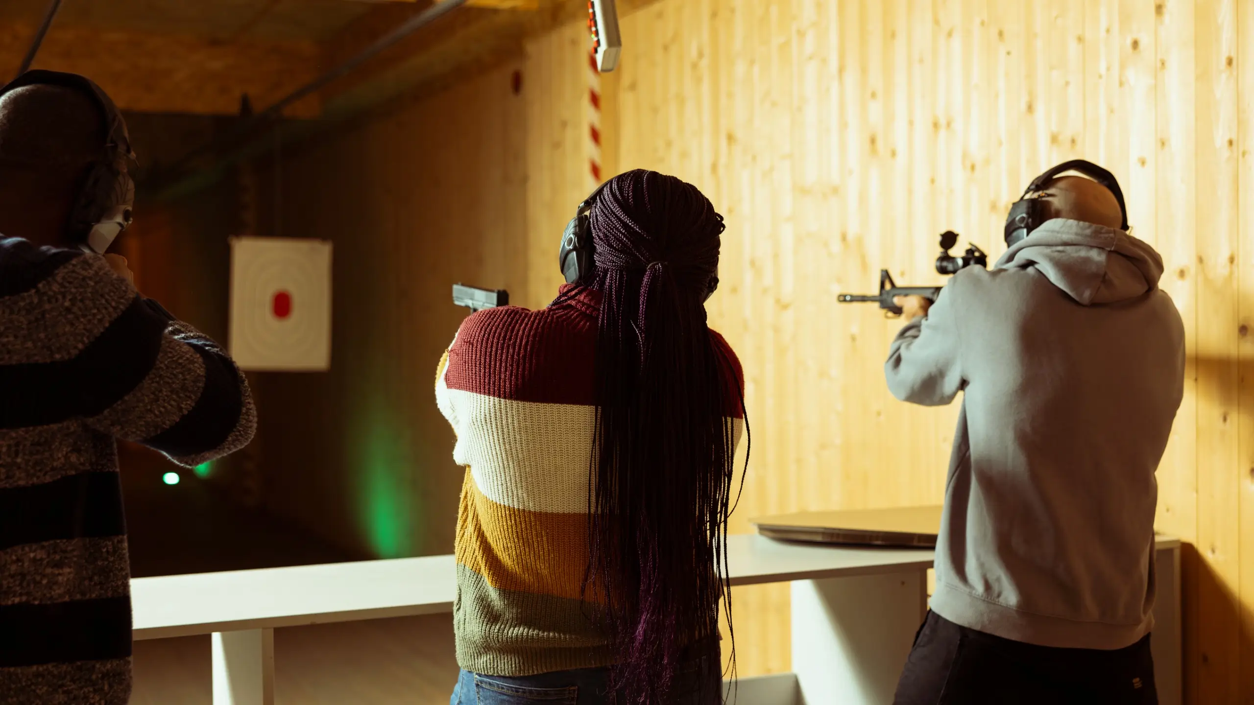 Three people shooting at an indoor range from behind, one with a handgun and one with a scoped rifle, all wearing hearing protection.