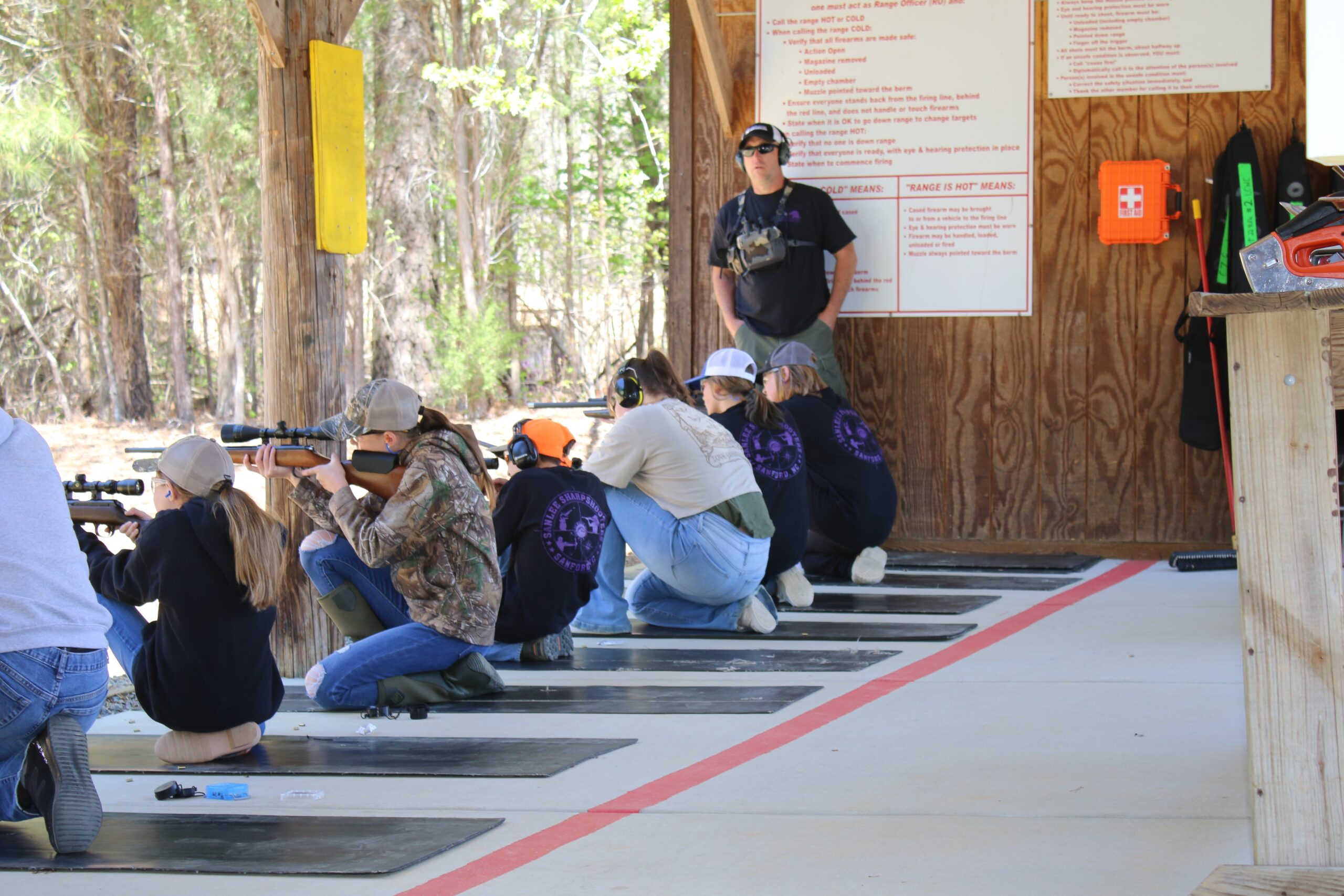 Young shooters kneeling at an outdoor covered firing line with scoped rifles, supervised by a standing range safety officer, with range rules posted on the wall behind them.