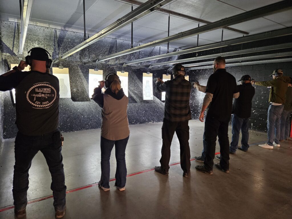 Group of shooters with hearing protection firing handguns at paper silhouette targets on an indoor range firing line.
