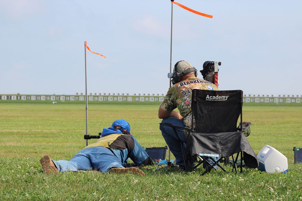 Prone shooter and seated spotter working together at an outdoor long-range shooting competition, with numbered target boards visible downrange and wind flags indicating conditions.