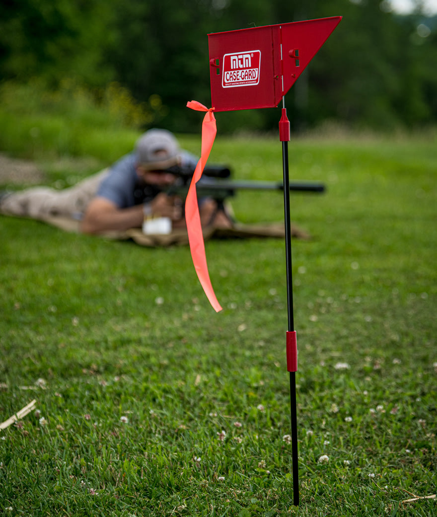 MTM Case-Gard wind flag with orange streamer staked in grass at a shooting range, with a prone shooter aiming a scoped bolt-action rifle in the background.