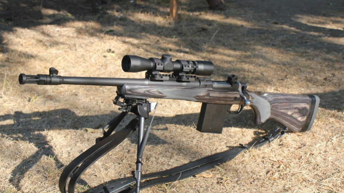 Bolt-action rifle with laminate thumbhole stock, detachable box magazine, muzzle brake, bipod, and riflescope, resting on dry ground.