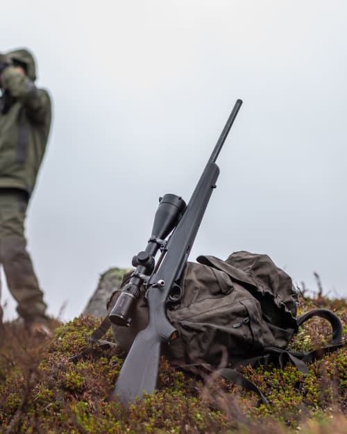 Bolt-action rifle with scope resting on a hunting pack on a moorland hillside, hunter glassing in the background.