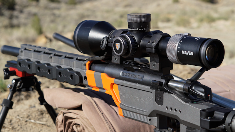 Close-up of a Bergara precision bolt-action rifle with orange and gray chassis, mounted with a Maven riflescope, resting on a bipod outdoors.