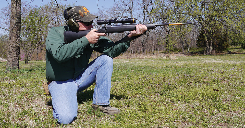 Man kneeling and aiming a scoped bolt-action rifle outdoors in a grassy field with trees in the background.