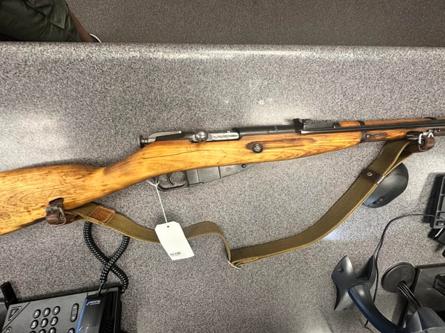 Mosin Nagant bolt-action rifle with wood stock, olive drab sling, and a white price tag, laid flat on a gray surface.