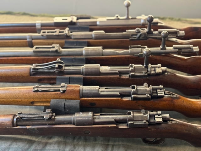 Multiple military surplus bolt-action rifles with wood stocks laid side by side on a green surface, showing receivers and bolt handles of various configurations.