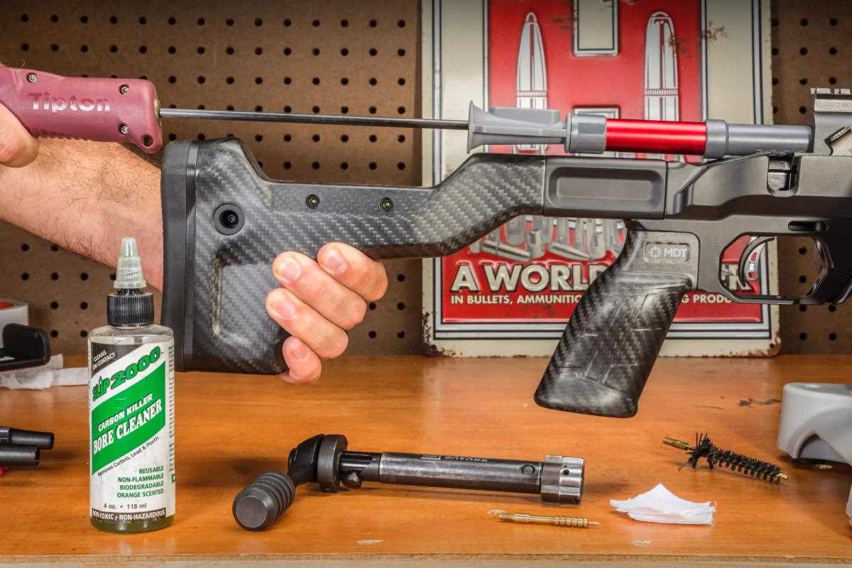 Bolt-action rifle in MDT chassis held at a workbench while a Tipton cleaning rod is run through the bore, with the bolt removed and CLP 2000 bore cleaner and cleaning brushes on the bench.