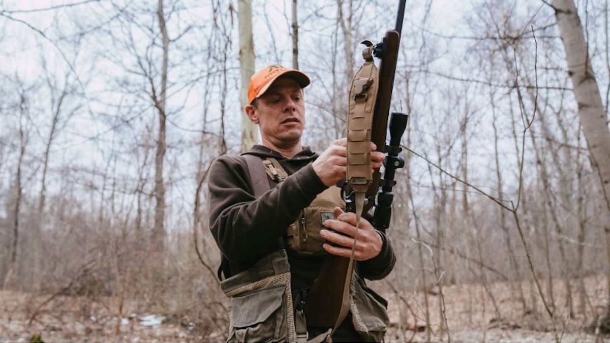 Hunter in blaze orange cap examining the barrel of a scoped bolt-action rifle in a bare winter woodland.