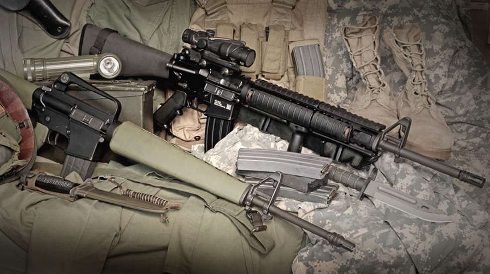 AR-15 rifle with 20-inch barrel and ACOG optic laid on military gear alongside magazines, a bayonet, and a stripped lower receiver.