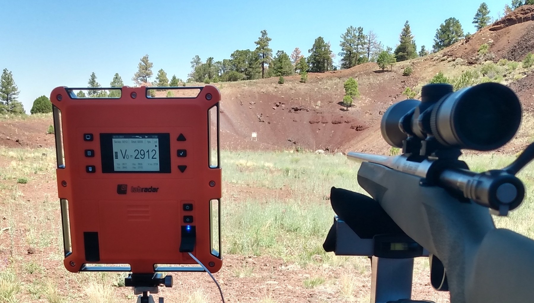 LabRadar Doppler chronograph on a tripod displaying a muzzle velocity of 2912 fps, with a scoped bolt-action rifle visible to the right, set up at an outdoor shooting range.