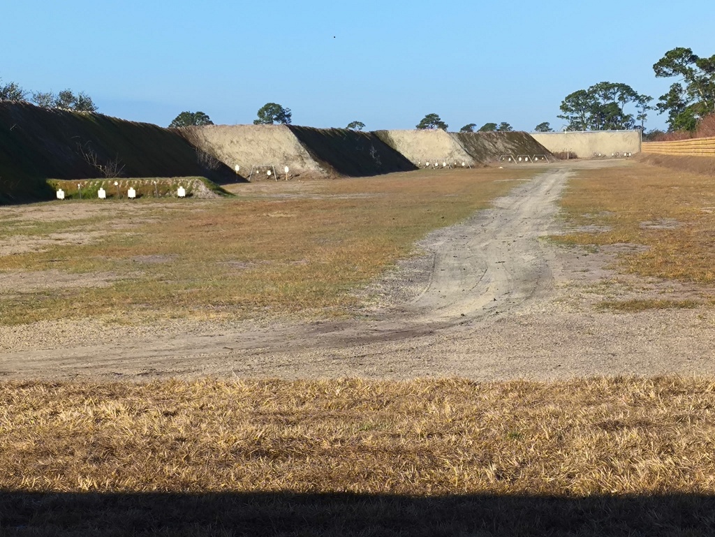 Outdoor shooting range with multiple earthen backstop berms and white paper silhouette targets at varying distances, viewed from the firing line.