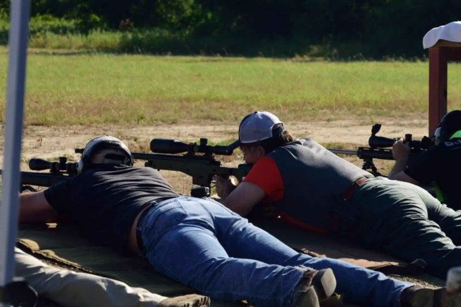 Two shooters lying prone at an outdoor shooting range, each behind a scoped bolt-action rifle on a bipod, with a third shooter visible at right.