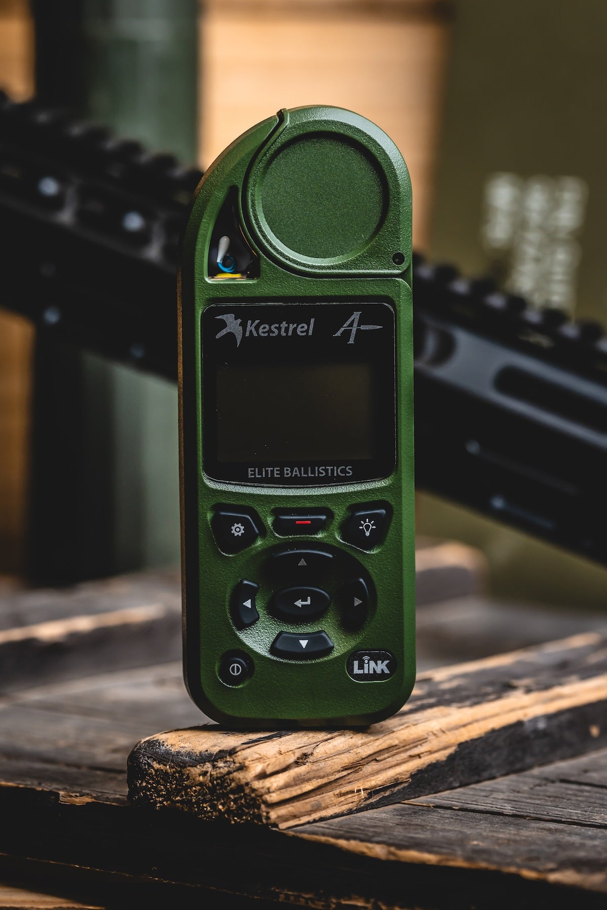 Kestrel Elite Ballistics weather meter in olive drab green with LiNK button, standing upright on a wooden surface at a shooting range.