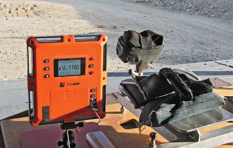 Orange LabRadar Doppler radar chronograph on a tripod displaying V0 equals 1160 on its screen, sitting on a shooting bench next to sandbags at an outdoor range.