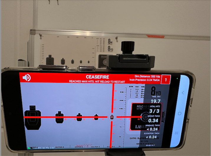 Smartphone mounted on a laser training simulator displaying a ceasefire screen with group size and hit data from a simulated 500-yard shooting exercise.
