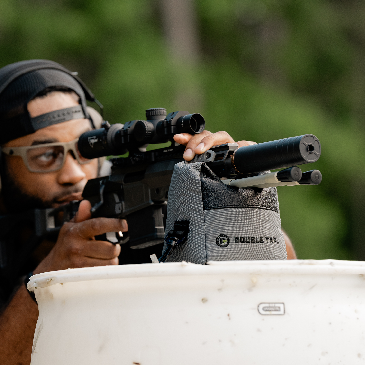 Shooter aiming a suppressed bolt-action rifle through a riflescope, supported on a Double Tap shooting bag rested on a barrel, wearing ear protection and safety glasses.