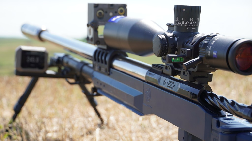 Close-up of a Warwick Tactical precision bolt-action rifle on a bipod, fitted with a large Zeiss riflescope and a bubble level on the scope mount, set against an open field.