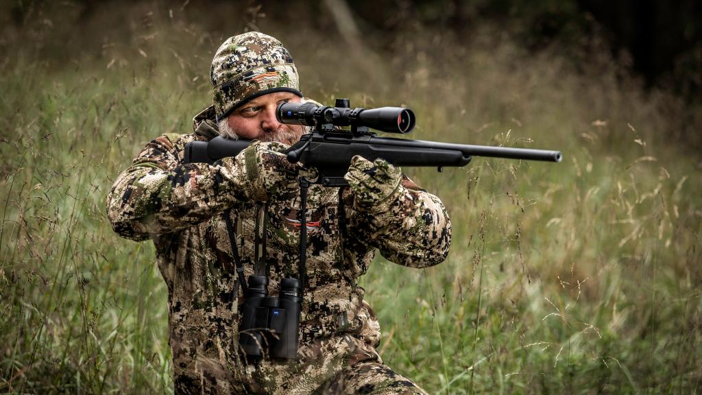 Hunter in camouflage kneeling in tall grass, aiming a bolt-action rifle mounted with a riflescope.