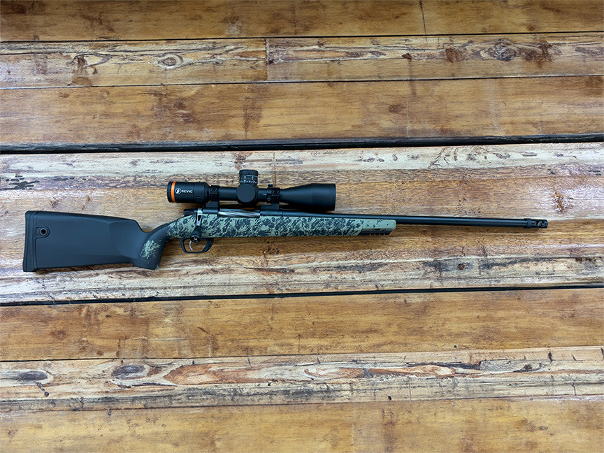 Bolt-action hunting rifle with green camouflage stock lying on a wooden surface, topped with a Revic Acura RS25i riflescope.