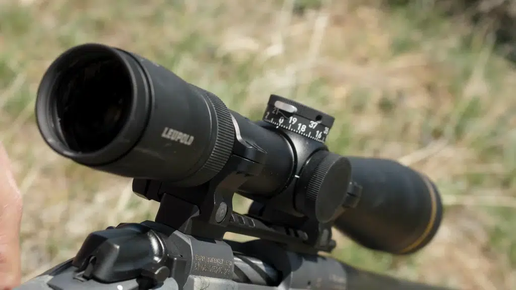 Close-up of a Leupold riflescope mounted on a rifle receiver, with the objective lens and magnification ring in focus against a blurred grassy background.