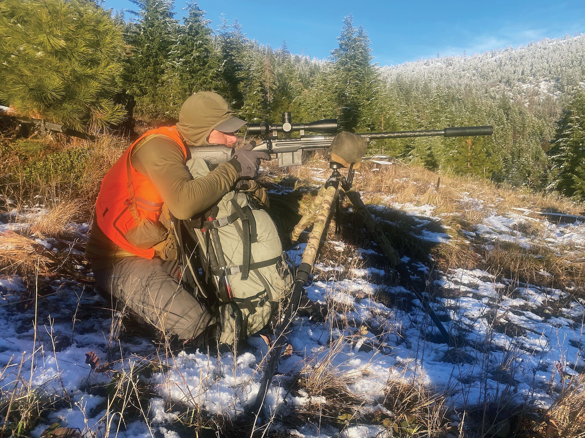 Hunter in blaze orange vest kneeling in snowy mountain terrain, aiming a suppressed bolt-action rifle mounted on a tripod with a riflescope.
