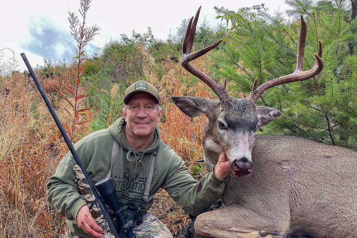 Hunter kneeling beside a harvested blacktail deer buck in dense autumn brush, with a scoped bolt-action rifle propped nearby.