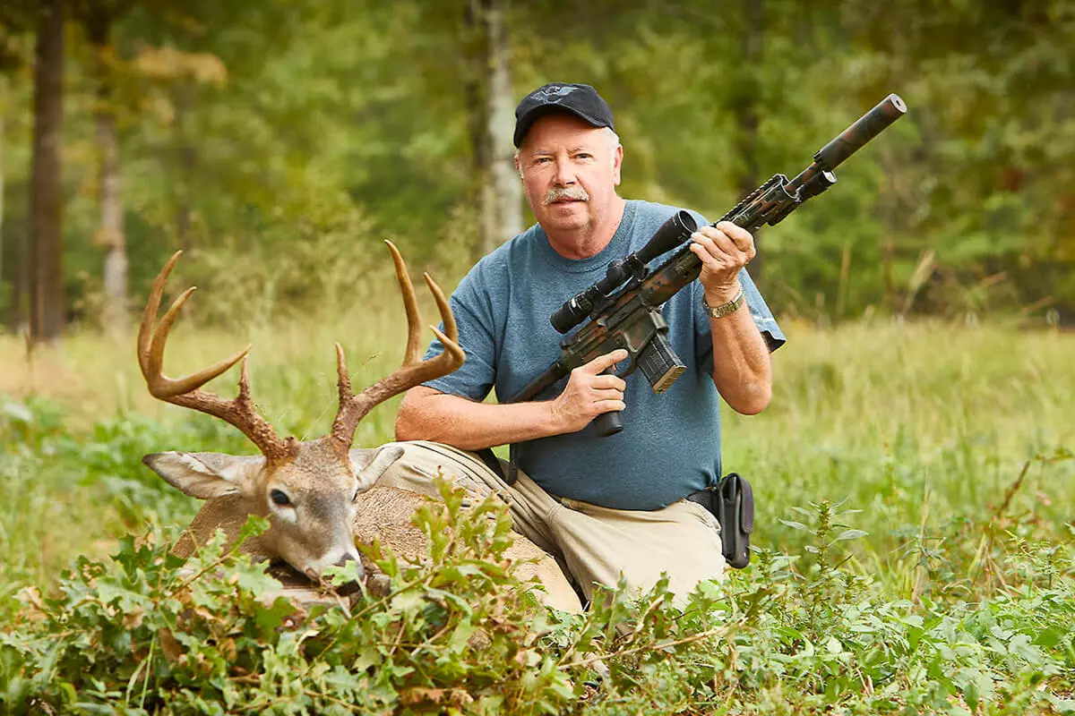 Hunter kneeling beside a harvested whitetail buck, holding a suppressed, scoped AR-style rifle in camouflage finish.