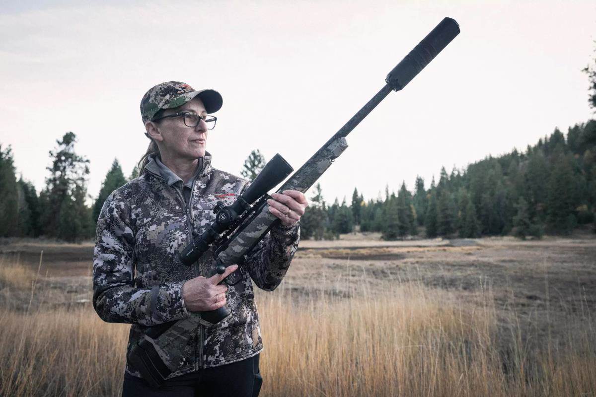 A woman in camouflage hunting gear holds a suppressed bolt-action rifle with a riflescope in an open field with trees in the background.
