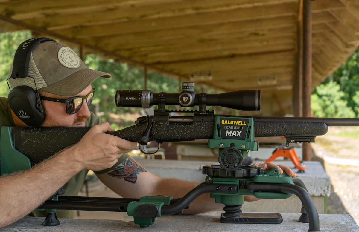 Shooter prone at an outdoor range bench, aiming a bolt-action rifle topped with a Maven scope, rested in a Caldwell Lead Sled MAX shooting rest.