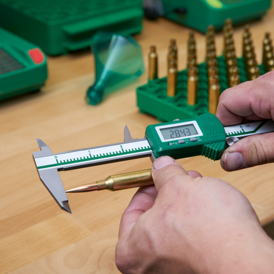 Hands using a green digital caliper to measure the overall length of a rifle cartridge at a reloading bench with a tray of loaded rounds in the background.