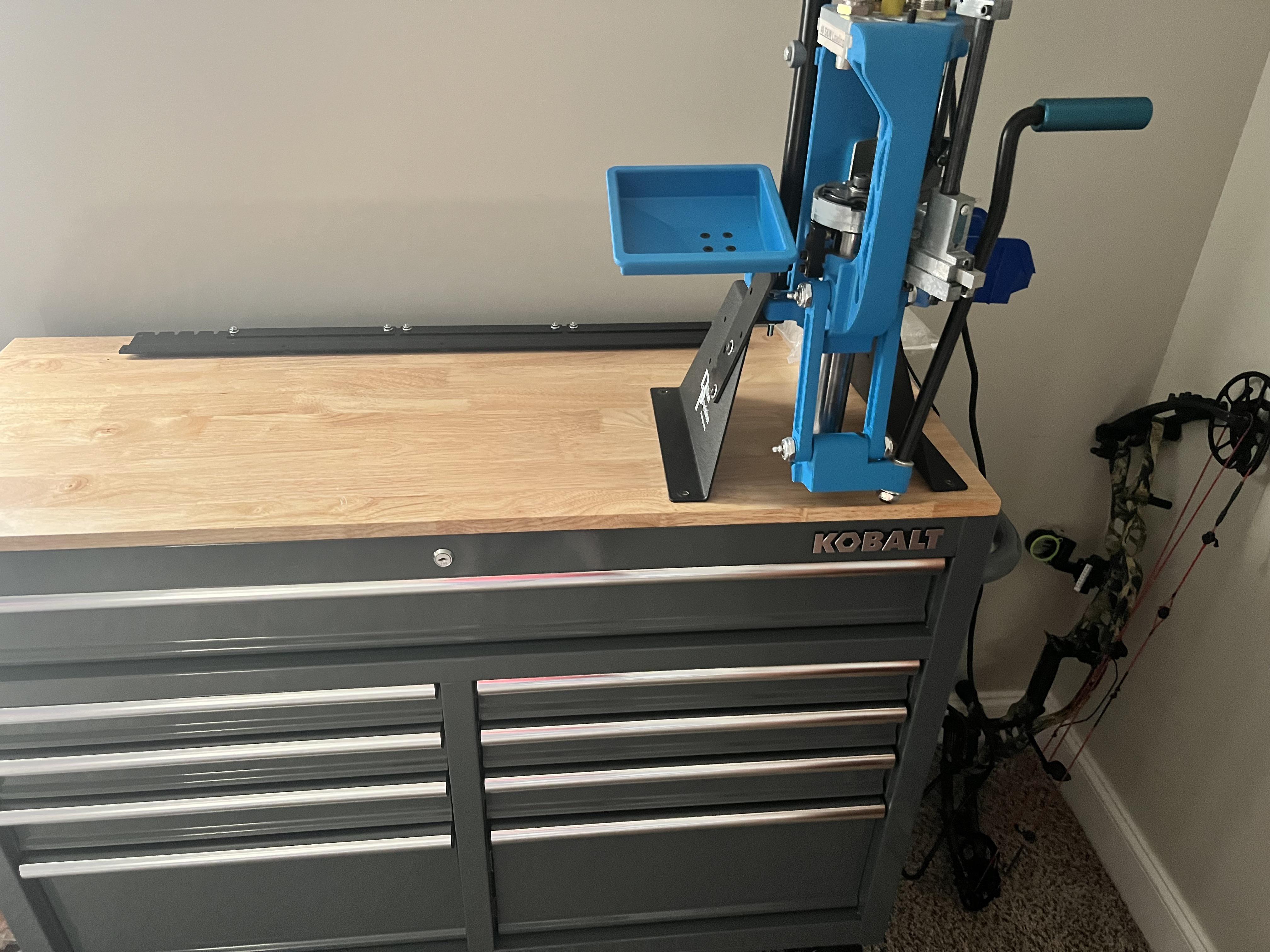 Blue reloading press bolted to the wooden top of a Kobalt tool chest in a home setup, with a compound bow leaning against the wall in the background.