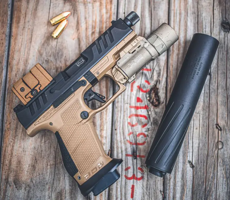 Heckler and Koch P30 pistol in flat dark earth finish with a weapon light attached, next to a detached black suppressor, on a weathered wood surface with loose 9mm ammunition nearby.