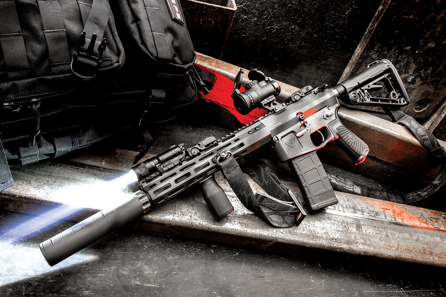 Short-barreled AR-style rifle with suppressor, red dot sight, and mounted flashlight resting on a metal surface with a tactical bag in the background.