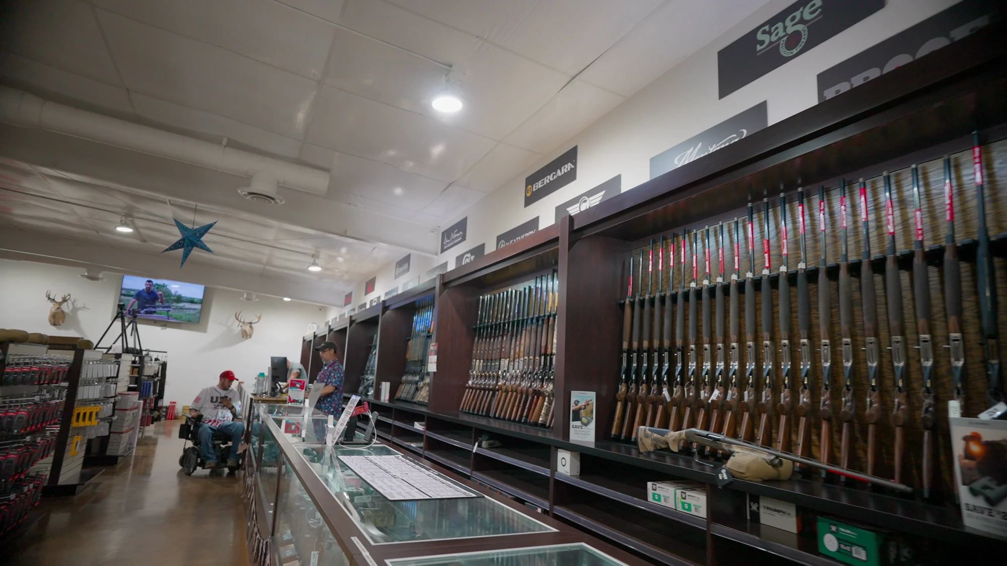 Interior of a gun store showing long gun racks filled with rifles and shotguns, glass display cases, and brand signage including Bergara and Sage, with customers browsing.