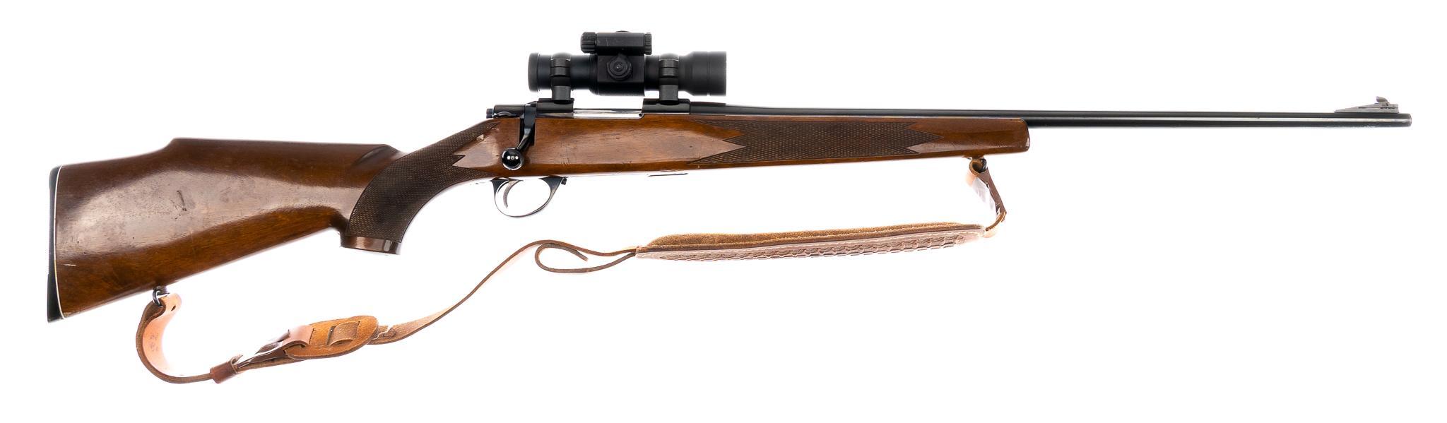 Blued bolt-action rifle with walnut stock, leather sling, and compact riflescope mounted in low rings, shown on white background.