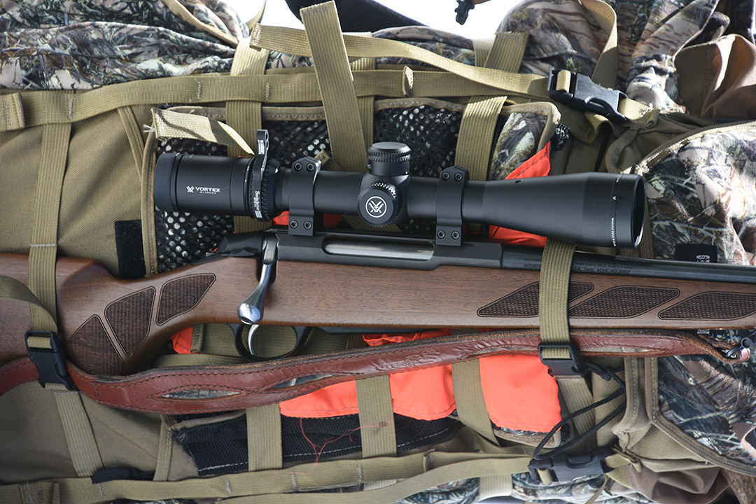 Sako bolt-action rifle with walnut stock and Vortex riflescope mounted in rings, resting on hunting packs and gear.