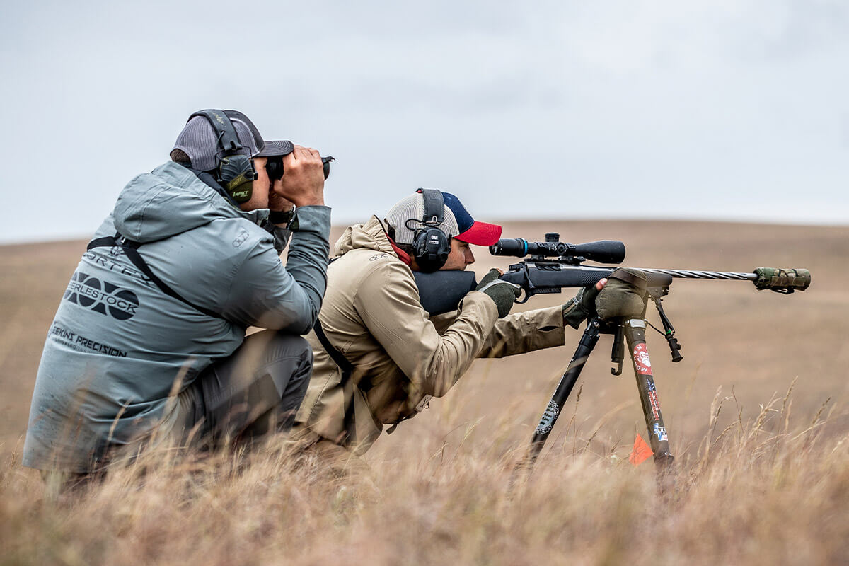 Two shooters in a field, one aiming a suppressed bolt-action rifle on a tripod, the other spotting with binoculars.